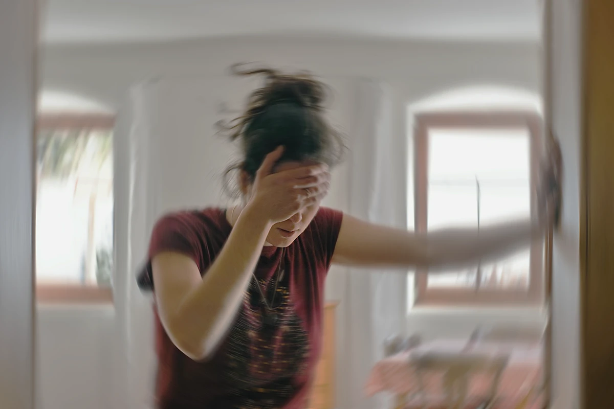 Blurred image of a woman holding her forehead and struggling to stay balanced in a bright room.