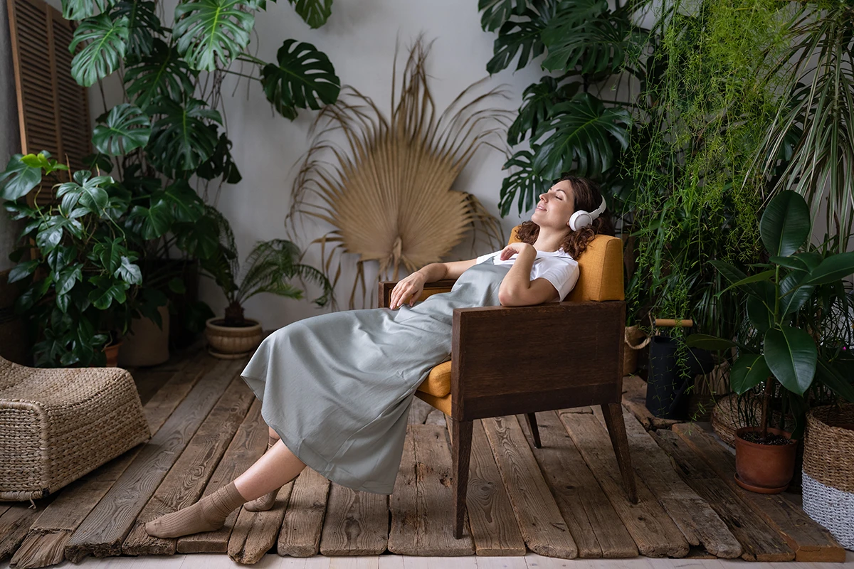 Woman relaxing in a chair with headphones, surrounded by lush green indoor plants.